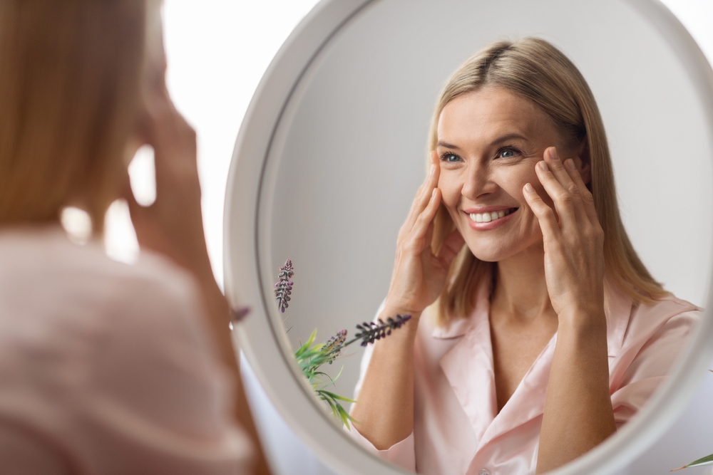 A smiling woman looking in the mirror, touching her face, considering skin rejuvenation options like Ulthera and facelift.