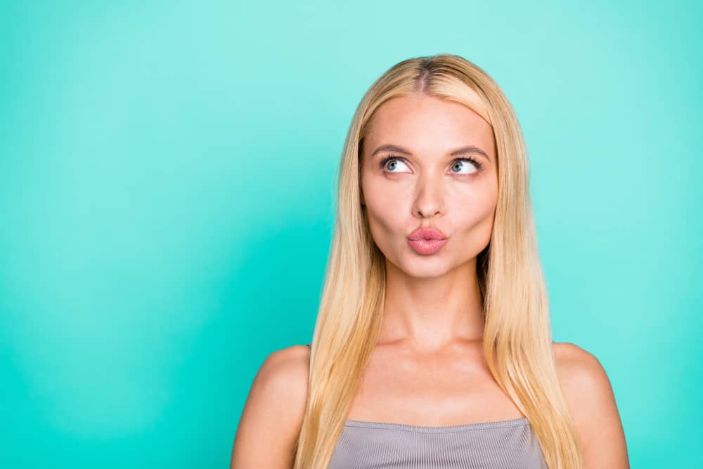 Woman with blonde hair making a pouting face, showcasing fuller lips after a lip augmentation procedure against a turquoise background.