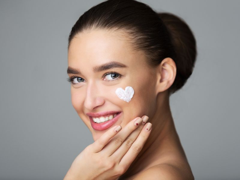 Smiling woman with glowing skin and a heart-shaped cream spot on her cheek, highlighting the benefits of VI Peels for youthful skin.